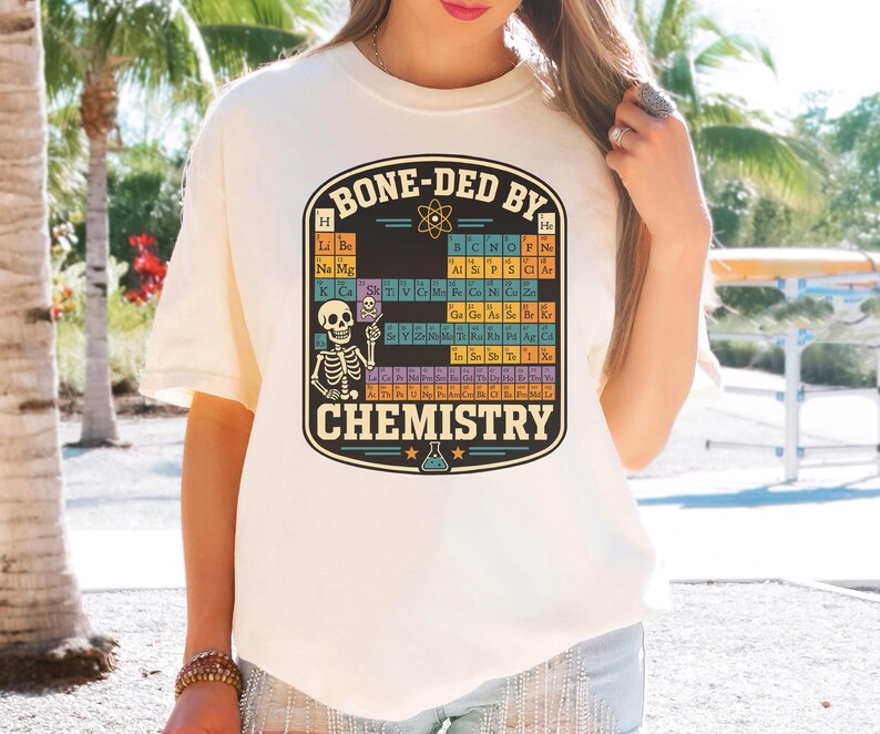 May include: A cream-colored t-shirt featuring a graphic print of a periodic table of elements with a skeleton pointing at a skull icon. The text BONE-DED BY CHEMISTRY is printed in a bold, dark font above and below the colorful table design.