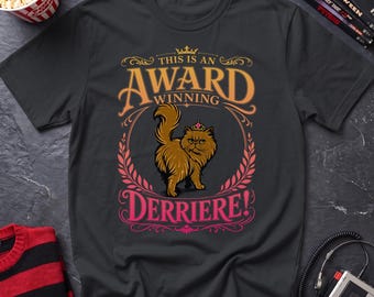 May include: A black t-shirt featuring a graphic of a fluffy brown cat wearing a small crown. The text reads "THIS IS AN AWARD WINNING DERRIERE!" in stylized orange and pink lettering, framed by decorative flourishes and a laurel wreath design.