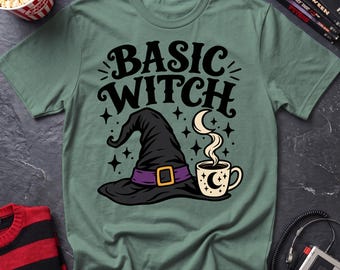 May include: A sage green short-sleeve t-shirt featuring a black graphic print that reads "BASIC WITCH" in stylized lettering. Below the text is a black witch hat with a purple band and a gold buckle, alongside a coffee mug with a crescent moon.