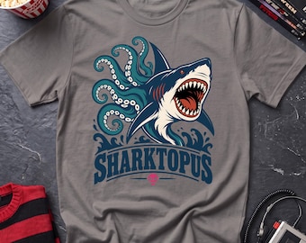 May include: A grey short-sleeved t-shirt featuring a graphic illustration of a shark with octopus tentacles. The design includes the text SHARKTOPUS in bold blue letters above a small pink skull icon, with the creature emerging from blue waves.