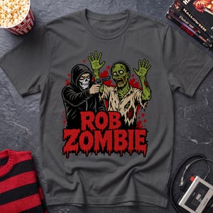 May include: A charcoal gray t-shirt featuring a graphic print of a hooded figure with a skull mask pointing a handgun at a green zombie with raised hands. The text "ROB ZOMBIE" appears in large, dripping red letters below the figures.