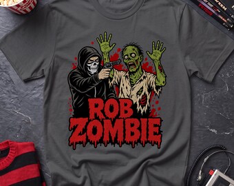 May include: A charcoal gray t-shirt featuring a graphic print of a hooded figure with a skull mask pointing a handgun at a green zombie with raised hands. The text "ROB ZOMBIE" appears in large, dripping red letters below the figures.