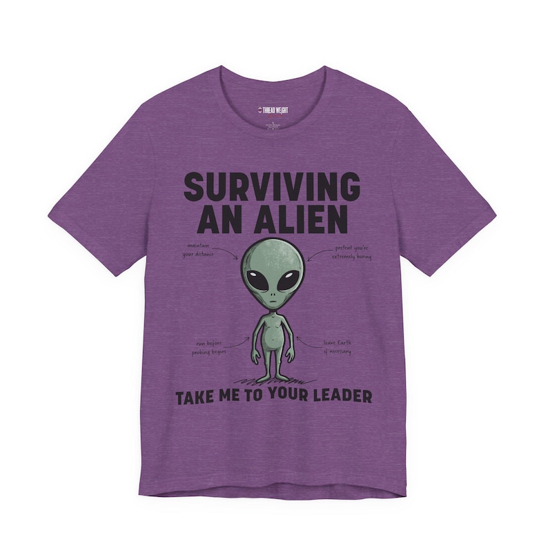 May include: A heather purple short-sleeve t-shirt featuring a graphic of a grey alien. The text reads: SURVIVING AN ALIEN, TAKE ME TO YOUR LEADER, and includes small labels pointing to the alien: maintain your distance, pretend you're extremely boring, flee before probing begins, and leave Earth if necessary.