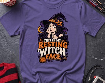May include: A purple short-sleeve t-shirt featuring a graphic of a witch wearing a hat with an orange flower. The text reads, "THIS IS MY RESTING WITCH FACE," accompanied by a crescent moon, bats, and a broomstick illustration in orange and white.