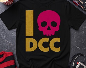 May include: A black short-sleeve t-shirt featuring a graphic design with a large vertical gold letter I, a bright pink skull icon, and the gold letters DCC positioned below the skull. The design has a distressed, textured appearance.