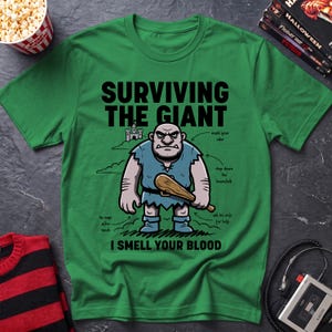 May include: A green t-shirt featuring a graphic of a cartoon giant holding a wooden club. The text reads "SURVIVING THE GIANT" and "I SMELL YOUR BLOOD," with smaller labels pointing to the giant, including "mask your oder," "chop down the beanstalk," "ask his wife for help," and "he naps after lunch."