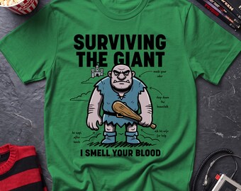 May include: A green t-shirt featuring a graphic of a cartoon giant holding a wooden club. The text reads "SURVIVING THE GIANT" and "I SMELL YOUR BLOOD," with smaller labels pointing to the giant, including "mask your oder," "chop down the beanstalk," "ask his wife for help," and "he naps after lunch."