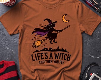 May include: A rust-colored short-sleeve t-shirt featuring a graphic of a witch flying on a broomstick against a crescent moon. Below the illustration, the text reads "LIFE'S A WITCH AND THEN YOU FLY" in bold black capital letters.
