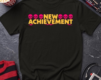 May include: A black short-sleeved t-shirt featuring the text NEW ACHIEVEMENT in yellow block letters. The design includes six small pink skull icons positioned above the word NEW and below the word ACHIEVEMENT, with pink lightning bolt accents.