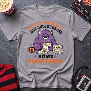 May include: A grey short-sleeve t-shirt featuring a purple one-eyed monster graphic. The monster is writing on papers with a pencil next to a mug. The text reads: NOT ALL MONSTERS LIVE UNDER THE BED SOME GRADE PAPERS in orange and black lettering.