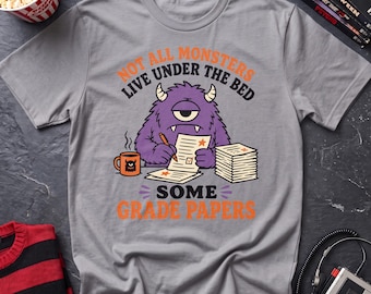 May include: A grey short-sleeve t-shirt featuring a purple one-eyed monster graphic. The monster is writing on papers with a pencil next to a mug. The text reads: NOT ALL MONSTERS LIVE UNDER THE BED SOME GRADE PAPERS in orange and black lettering.
