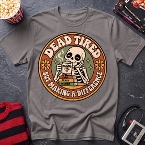 May include: A grey short-sleeved t-shirt featuring a circular graphic of a skeleton drinking from a mug labeled "TEACHER FUEL" with a heart. Below the mug are books labeled "PLAN," "TEACH," and "INSPIRE." The text reads "DEAD TIRED BUT MAKING A DIFFERENCE."