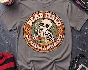 May include: A grey short-sleeved t-shirt featuring a circular graphic of a skeleton drinking from a mug labeled "TEACHER FUEL" with a heart. Below the mug are books labeled "PLAN," "TEACH," and "INSPIRE." The text reads "DEAD TIRED BUT MAKING A DIFFERENCE."