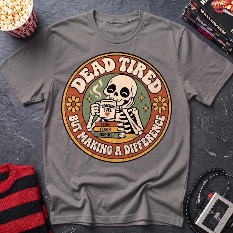 May include: A grey short-sleeved t-shirt featuring a circular graphic of a skeleton drinking from a mug labeled "TEACHER FUEL" with a heart. Below the mug are books labeled "PLAN," "TEACH," and "INSPIRE." The text reads "DEAD TIRED BUT MAKING A DIFFERENCE."