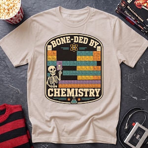 May include: A beige short-sleeve t-shirt featuring a graphic of a periodic table with a skeleton pointing to the element Sk. The text BONE-DED BY CHEMISTRY is printed in a bold font above and below the table, with an atom and flask icon.