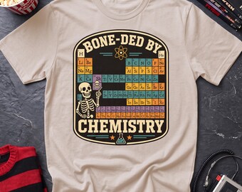 May include: A beige short-sleeve t-shirt featuring a graphic of a periodic table with a skeleton pointing to the element Sk. The text BONE-DED BY CHEMISTRY is printed in a bold font above and below the table, with an atom and flask icon.