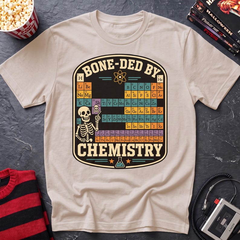 May include: A beige short-sleeve t-shirt featuring a graphic of a periodic table with a skeleton pointing to the element Sk. The text BONE-DED BY CHEMISTRY is printed in a bold font above and below the table, with an atom and flask icon.