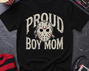May include: A black short-sleeved t-shirt featuring a graphic print of a white hockey mask with red accents. The text PROUD BOY MOM is printed in a distressed white font arched above and below the mask, which has a cracked texture.