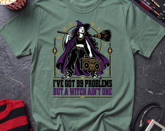 May include: A sage green t-shirt featuring a graphic of a witch wearing a purple cape, black crop top, and pants, holding a broomstick and a boombox. The text reads, I'VE GOT 99 PROBLEMS BUT A WITCH AIN'T ONE, surrounded by stars and geometric shapes.