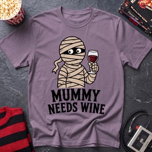 May include: A mauve short-sleeved t-shirt featuring a cartoon illustration of a mummy holding a glass of red wine. Below the graphic, the text reads "MUMMY NEEDS WINE" in bold black lettering. The shirt has a crew neckline and a standard fit.
