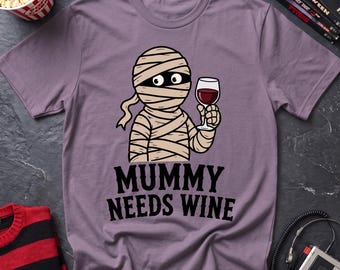 May include: A mauve short-sleeved t-shirt featuring a cartoon illustration of a mummy holding a glass of red wine. Below the graphic, the text reads "MUMMY NEEDS WINE" in bold black lettering. The shirt has a crew neckline and a standard fit.