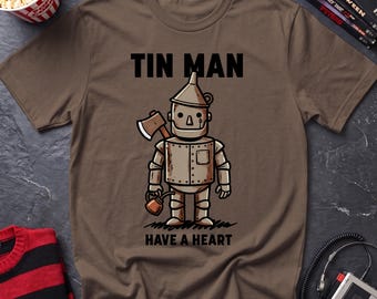 May include: A brown short-sleeved t-shirt featuring a graphic of the Tin Man character holding an axe and an oil can. The text "TIN MAN" is printed above the character, and the text "HAVE A HEART" is printed below the character in black ink.