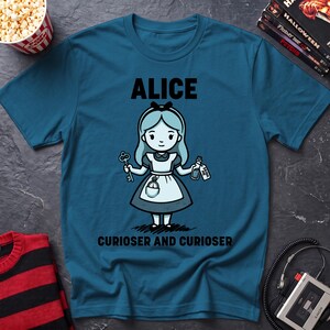 May include: A teal blue t-shirt featuring a cartoon illustration of Alice holding a key and a bottle with a tag that reads DRINK ME. The text ALICE appears above the character, and the text CURIOSER AND CURIOSER is printed below the character.