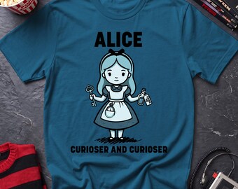 May include: A teal blue t-shirt featuring a cartoon illustration of Alice holding a key and a bottle with a tag that reads DRINK ME. The text ALICE appears above the character, and the text CURIOSER AND CURIOSER is printed below the character.