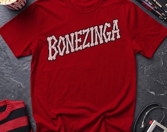 May include: A dark red short-sleeve t-shirt featuring the text BONEZINGA printed in a diagonal orientation across the chest. The letters are stylized to resemble light pink and white human bones with a dark outline, creating a graphic design.