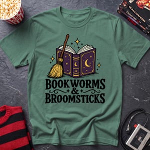 May include: A sage green t-shirt featuring a graphic of an open purple book with gold crescent moon and star details, accompanied by a straw broomstick. Below the illustration, the text reads "BOOKWORMS & BROOMSTICKS" in black lettering.