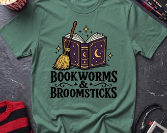 May include: A sage green t-shirt featuring a graphic of an open purple book with gold crescent moon and star details, accompanied by a straw broomstick. Below the illustration, the text reads "BOOKWORMS & BROOMSTICKS" in black lettering.