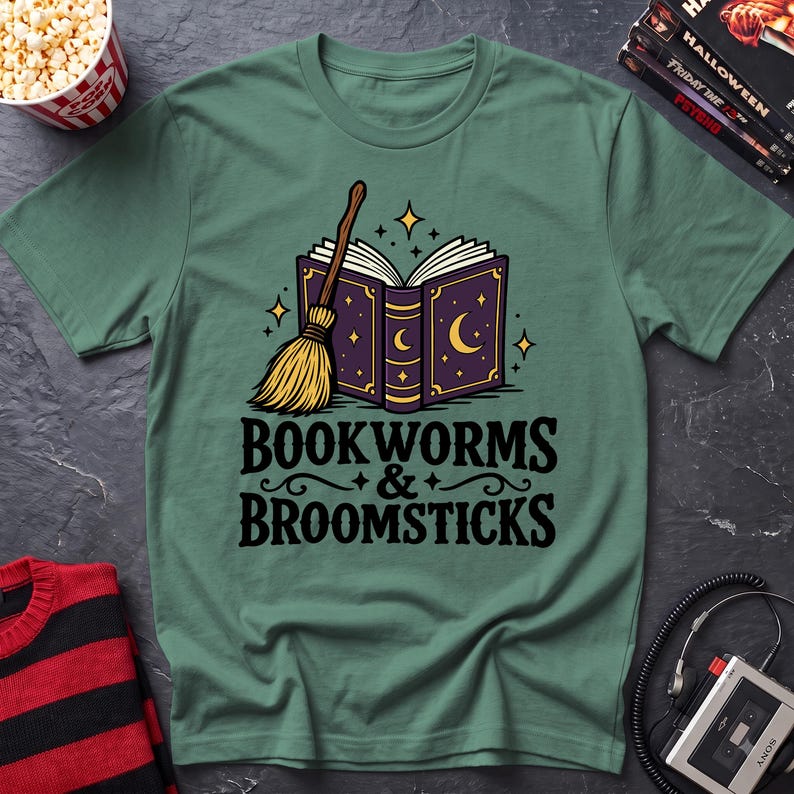 May include: A sage green t-shirt featuring a graphic of an open purple book with gold crescent moon and star details, accompanied by a straw broomstick. Below the illustration, the text reads "BOOKWORMS & BROOMSTICKS" in black lettering.