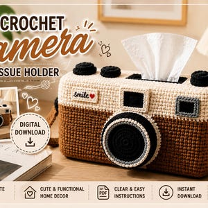 Könnte beinhalten: Ein gehäkelter Taschentuchhalter in Kameraform mit cremefarbenem Oberteil und braunem Unterteil sowie schwarzem Objektiv, Knöpfen und Trageriemen. Ein kleines weißes Etikett trägt die Aufschrift smile und ein rotes Herz.