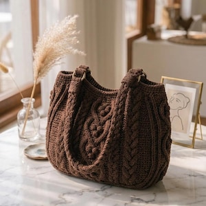 Puede incluir: Un bolso de ganchillo de color marrón oscuro con un patrón de punto de cable texturizado, detalles trenzados verticales y asas integradas unidas con anillos metálicos dorados. El bolso tiene una forma redondeada y estructurada, y un tejido grueso.