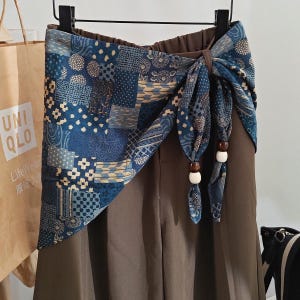 May include: A pair of solid brown trousers featuring an attached wrap-around waist sash. The sash is made of blue fabric with a patchwork pattern of geometric and floral motifs, tied at the side with dangling brown and white wooden beads.