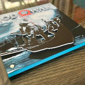 May include: A square, multi-layered wall art piece featuring the God of War logo and a dark silhouette of two figures in a boat on a blue and white icy background. The design has a raised, 3D effect with visible layers and silver metal fasteners.