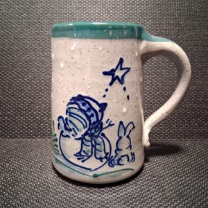 May include: A ceramic mug with a whimsical winter scene. The mug features a speckled white base, a teal rim, and a white handle. A blue illustration depicts a snowman, a star, and a rabbit. The mug is approximately 4 inches tall.