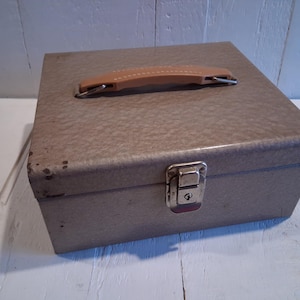 Puede incluir: Una caja metálica rectangular vintage con un acabado texturizado marrón claro. Cuenta con un asa de cuero marrón claro y un pestillo plateado con cerradura. La caja está sobre una superficie de madera blanca.