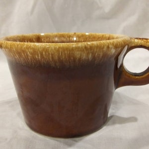 May include: A brown ceramic mug with a light brown glaze. The mug has a large handle and a wide, rounded shape.