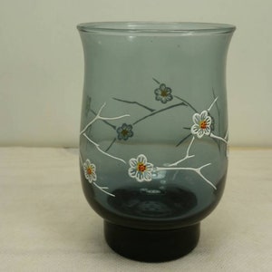 May include: A gray glass vase with a floral design. The vase has a wide base and a narrow neck. The flowers are white with yellow centers and are painted on the vase.