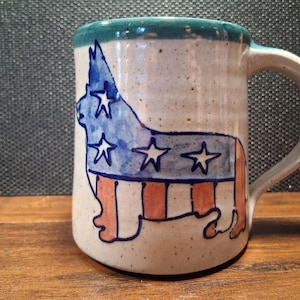 May include: A ceramic mug with a handle, featuring a patriotic design. The mug showcases a dog silhouette with a blue and white star design and red and white stripes, reminiscent of the American flag. The rim is teal.