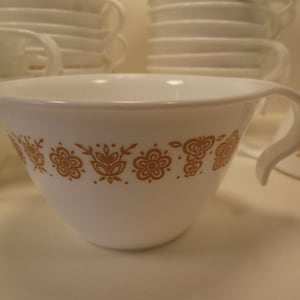 May include: White ceramic teacup with a curved handle and a floral pattern in shades of brown. Several other cups are stacked in the background. The teacup has a wide opening and a rounded base, suitable for hot beverages.