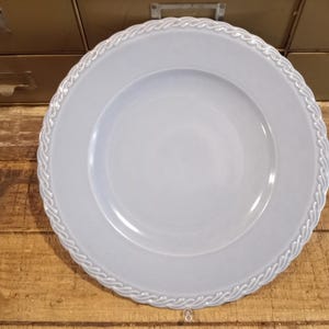 Könnte beinhalten: Ein hellblaues Keramik-Teller mit einem dekorativen, seilartigen Rand. Der Teller ist rund und hat eine leicht vertiefte Mitte. Der Teller liegt auf einer Holzoberfläche.