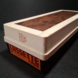 May include: A rectangular cassette case with a cream-colored body and a wood-grain patterned top. The case has an orange and black label that reads "CASSETTE CASE". The case is slightly worn.