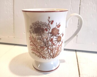 May include: White ceramic mug with a brown and red floral design. The mug has a handle and a pedestal base.