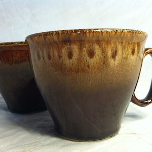 May include: Two brown ceramic mugs with a glazed finish. The mugs have a light brown top and a darker brown bottom. One mug has a handle.