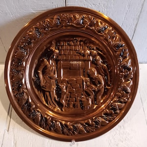 May include: A copper-colored decorative plate with a detailed relief scene of figures near a building. The plate has a raised border with a grape vine design. The plate is round and has a shiny finish.