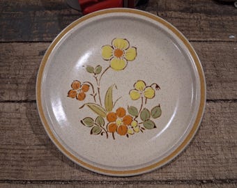 Puede incluir: Un plato redondo de cerámica con una base de color blanco roto moteado y un borde marrón. El centro presenta un diseño floral con flores amarillas y naranjas, hojas verdes y tallos. El plato tiene un acabado liso y esmaltado.