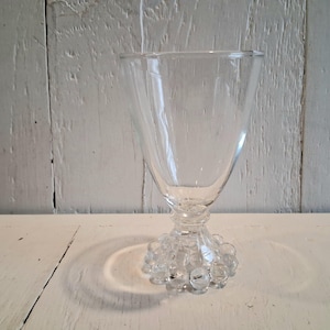 May include: Clear glass goblet with a wide, flared bowl and a stem adorned with small, round glass beads. The glass sits on a white wooden surface against a white, textured background.
