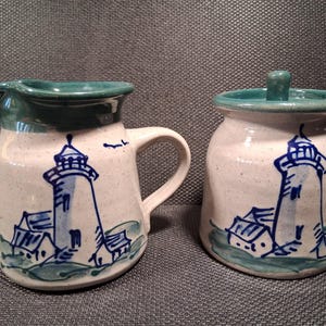 May include: A ceramic creamer pitcher and a lidded sugar bowl with a speckled off-white glaze. Both pieces feature a hand-painted blue lighthouse illustration with green accents on the rim and base. The pitcher has a handle and a pouring spout.